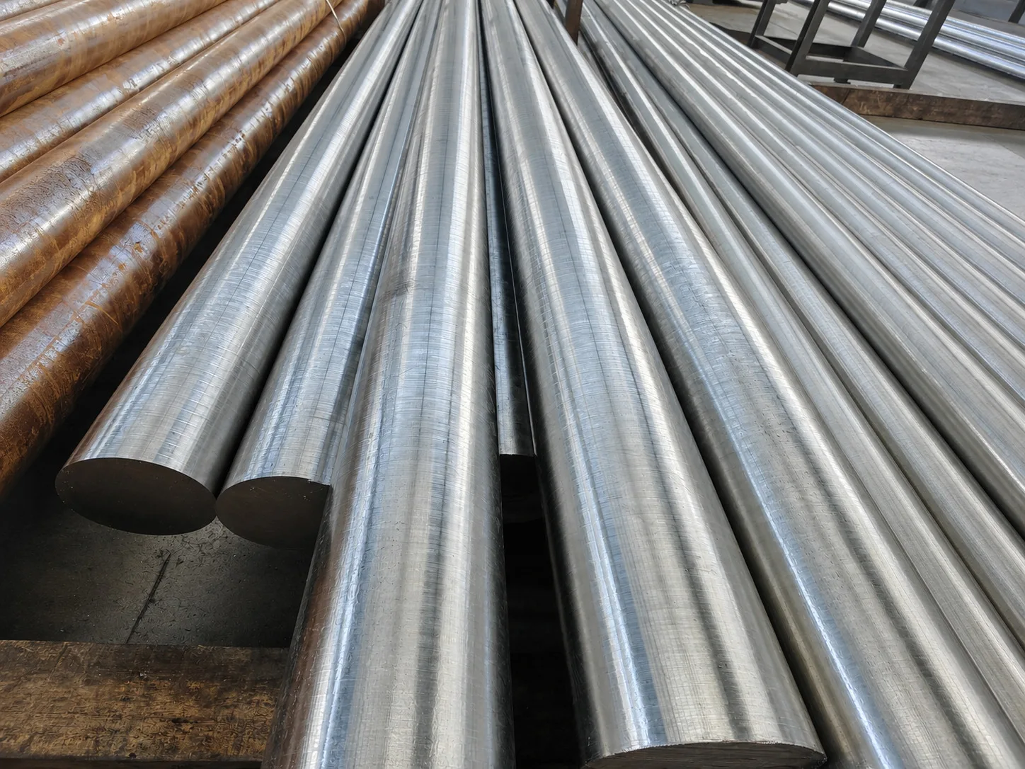 Rows of large cylindrical metal stock with dark and bright surfaces on workshop supports